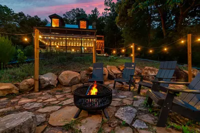 Cozy outdoor setting with a fire pit surrounded by black plastic chairs, illuminated by lights. Candles in the fire pit add to the warm and inviting ambiance. The scene includes a stone patio and a view of pine trees and mountains in the distance, creating a serene atmosphere perfect for a relaxing evening outdoors.