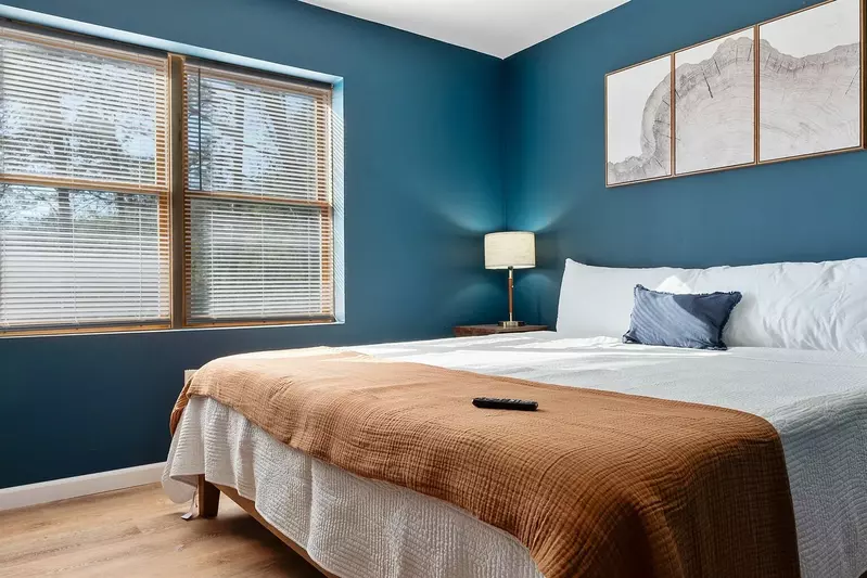 A cozy bedroom with a neatly made bed featuring a brown blanket and white pillows. The blue wall backdrop creates a calming ambiance, complemented by warm lighting from a lamp on the nightstand. Natural light streams in from a window, brightening the space. Decorative elements like framed pictures of mountain ranges adorn the walls. A remote control on the bed suggests a relaxing evening spent in this inviting bedroom.