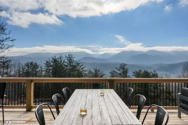 Cozy wooden deck with a large pine table surrounded by chairs of varying designs. One chair near the edge overlooks Great Smoky mountain in the distance. On the table, two glasses of beer invite relaxation and enjoyment of the view. The scene exudes a peaceful atmosphere, ideal for a mountain retreat or cabin getaway.