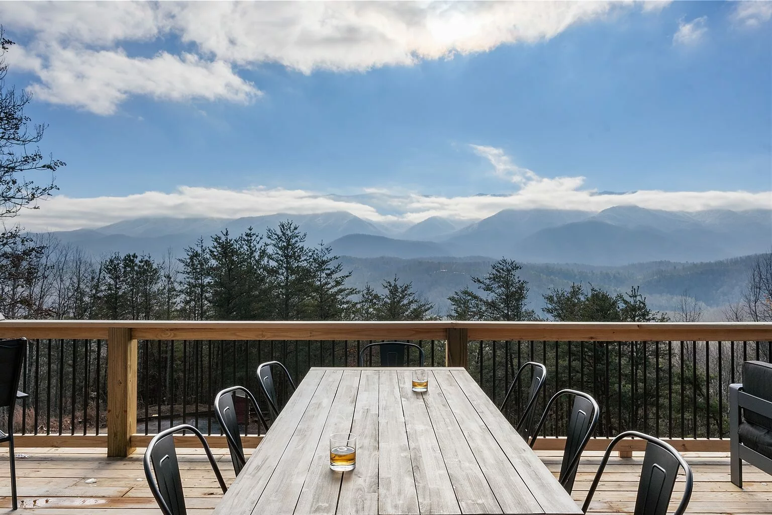 Cozy wooden deck with a large pine table surrounded by chairs of varying designs. One chair near the edge overlooks Great Smoky mountain in the distance. On the table, two glasses of beer invite relaxation and enjoyment of the view. The scene exudes a peaceful atmosphere, ideal for a mountain retreat or cabin getaway.