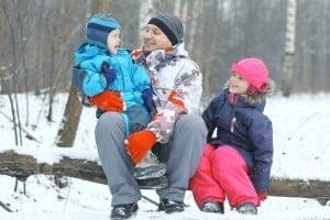 A dad and his young children sitting on a log in the woods during the winter.