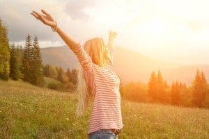 A young woman with her hands in the air as the sun shines behind the mountains.