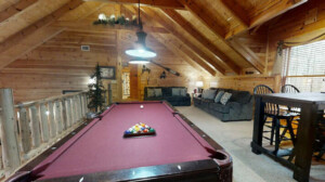 Upstairs game room with a pool table, 2 charcoal couches, 4 black bar stools at a black card table, loft ceiling