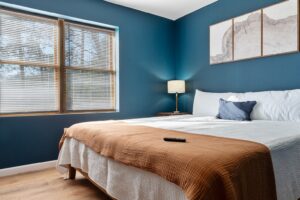 A cozy bedroom with a neatly made bed featuring a brown blanket and white pillows. The blue wall backdrop creates a calming ambiance, complemented by warm lighting from a lamp on the nightstand. Natural light streams in from a window, brightening the space. Decorative elements like framed pictures of mountain ranges adorn the walls. A remote control on the bed suggests a relaxing evening spent in this inviting bedroom.
