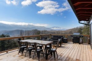 A spacious deck with a rustic wooden table and several metal chairs arranged neatly around it, set against a breathtaking backdrop of the majestic Great Smoky Mountains. The panoramic view includes towering mountains and a clear blue sky. The peaceful and serene scene is perfect for outdoor dining or relaxing, with comfortable seating options ideal for enjoying the fresh mountain air and picturesque surroundings.